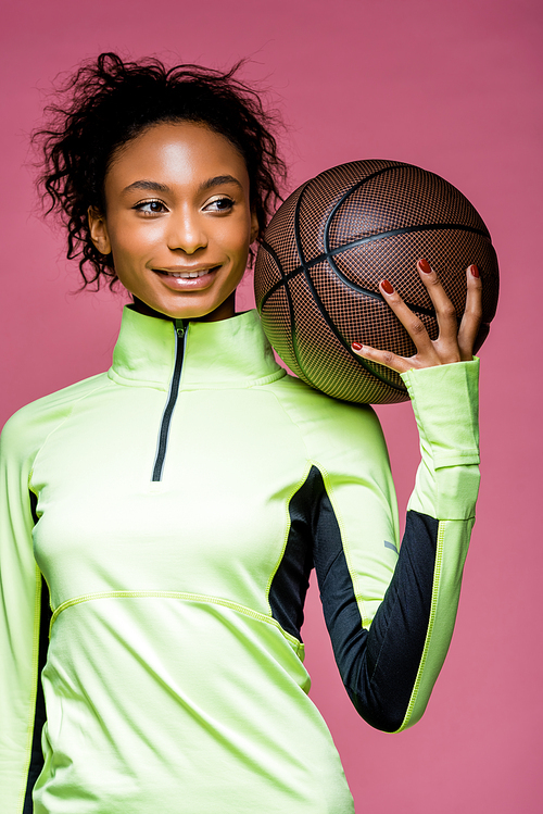 beautiful smiling african american sportswoman holding basketball isolated on pink