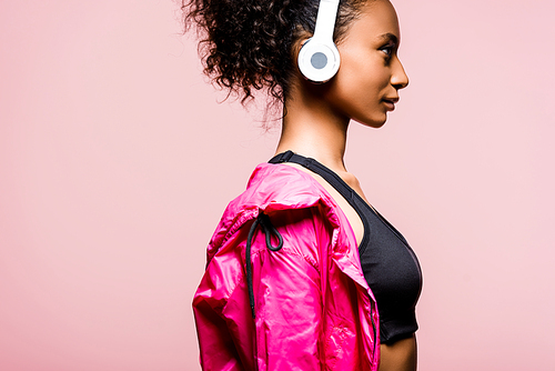 side view of beautiful african american sportswoman in windbreaker and headphones isolated on pink