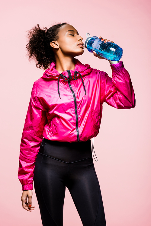 beautiful african american sportswoman drinking water from sport bottle isolated on pink