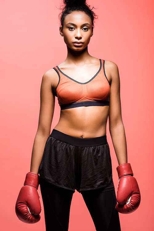 beautiful african american sportswoman in boxing gloves  and posing isolated on coral