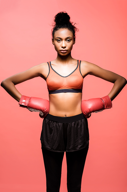 beautiful african american sportswoman in boxing gloves posing with hands akimbo and  isolated on coral