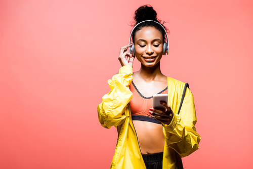 beautiful smiling african american sportswoman in headphones using smartphone isolated on coral with copy space