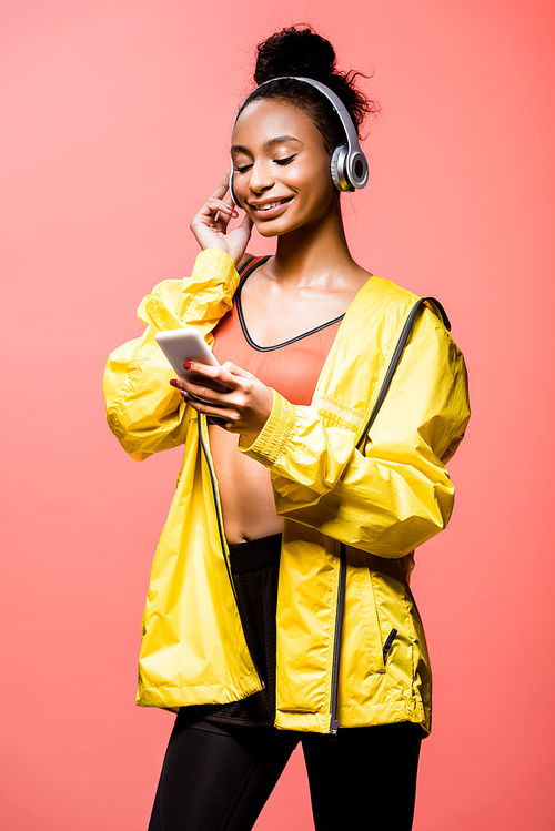 beautiful african american sportswoman in headphones smiling and using smartphone isolated on coral