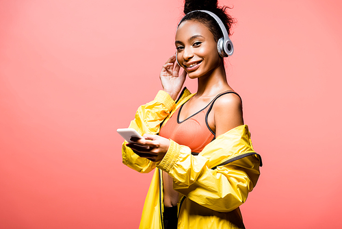 beautiful smiling african american sportswoman in headphones  and using smartphone isolated on coral