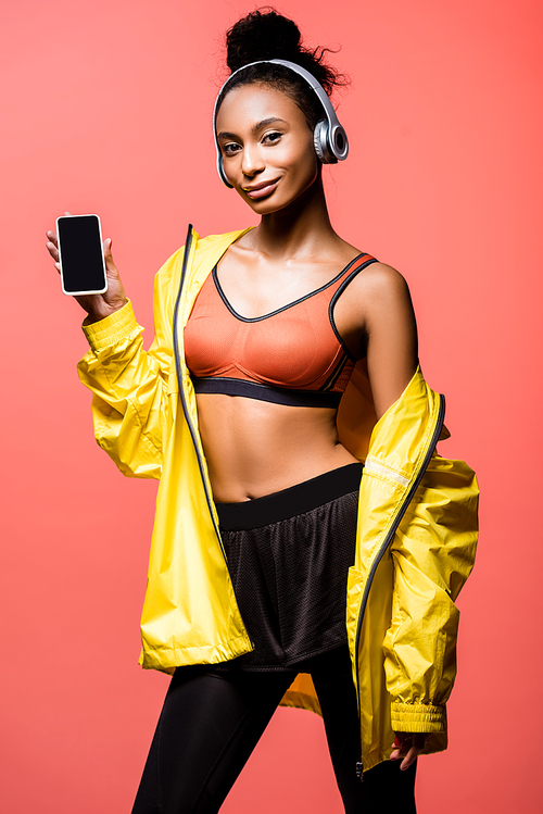 beautiful african american sportswoman in headphones  and presenting smartphone with blank screen isolated on coral