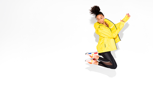 athletic african american sportswoman in yellow sports jacket jumping on white with copy space