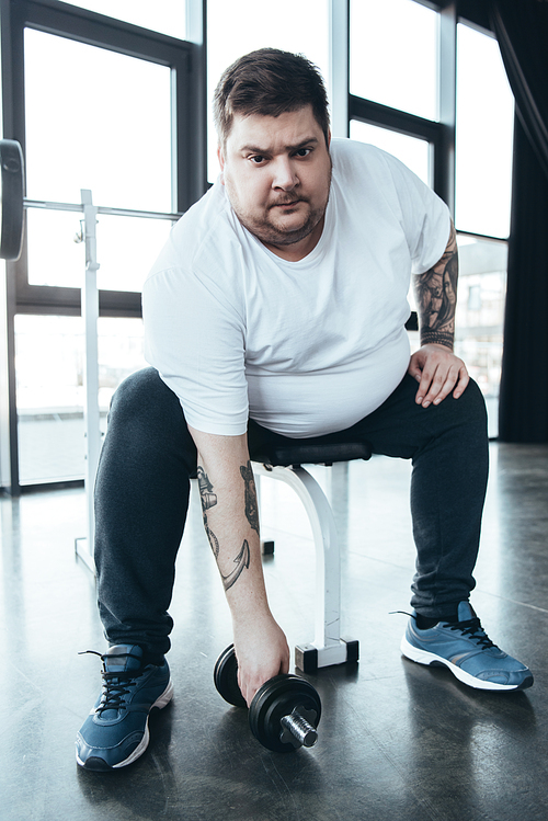 Overweight tattooed man Looking At Camera and exercising with dumbbell at sports center