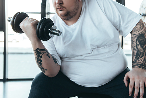 partial view of Overweight tattooed man in white t-shirt training with dumbbell at gym