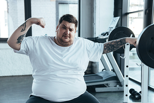 overweight tattooed man sitting on bench,  and showing muscles at gym