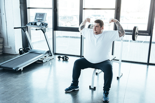 overweight tattooed man sitting on bench and showing muscles at gym
