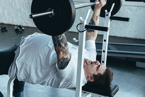 Overweight tattooed man in white t-shirt exercising with barbell at gym