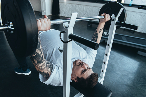 Obese tattooed man in white t-shirt training with barbell at gym
