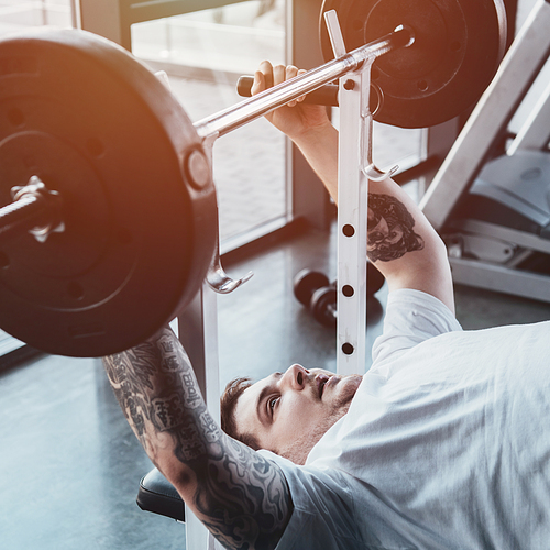 Overweight tattooed man training with barbell at gym with sunlight
