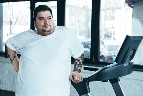 overweight tattooed man looking away near treadmill at sports center