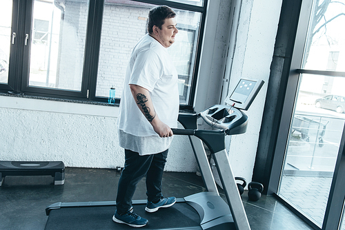 overweight tattooed man in white t-shirt running on treadmill at gym