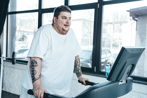 overweight tattooed man in white t-shirt running on treadmill at sports center