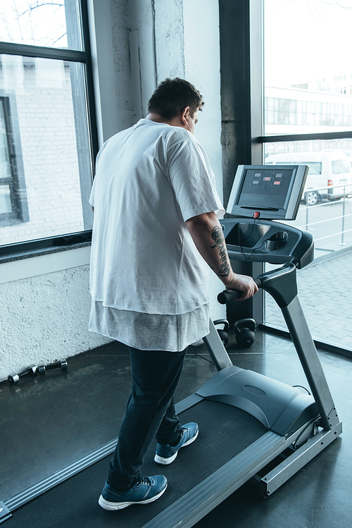 back view of overweight tattooed man running on treadmill at sports center