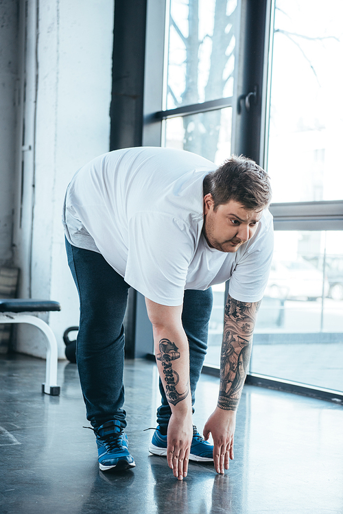 overweight tattooed man in white t-shirt doing stretching exercise at sports center