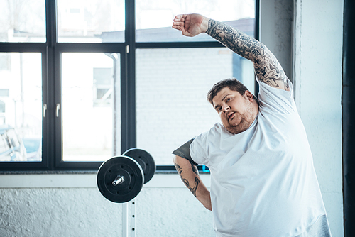 overweight tattooed man Looking At Camera and doing stretching exercise at gym