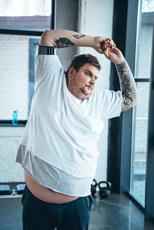 overweight tattooed man in white t-shirt stretching at sports center