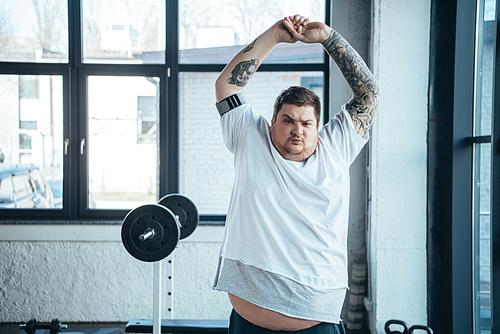 overweight tattooed man Looking At Camera and doing stretching exercise at gym