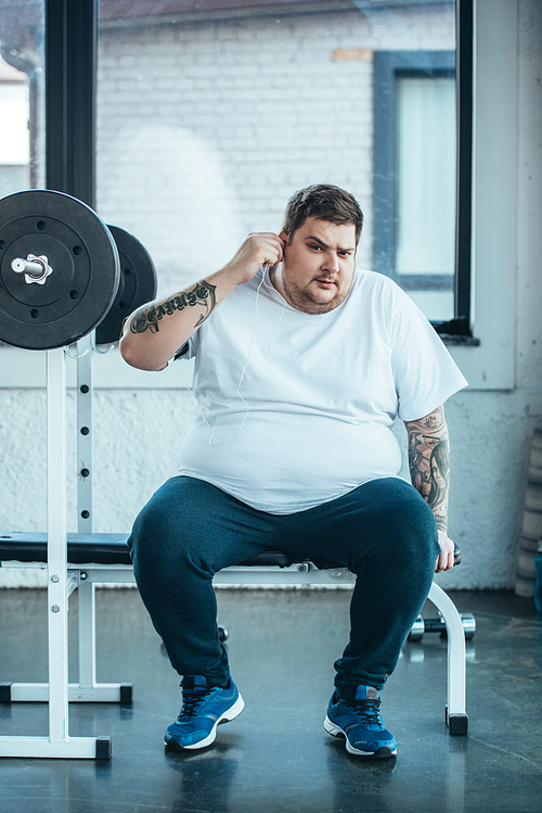 overweight tattooed man in earphones sitting on bench,  and listening music at gym