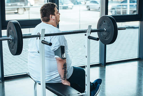 overweight man in earphones sitting on bench and listening music at gym