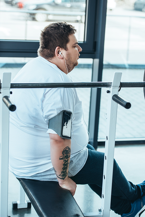 overweight man in earphones sitting on bench and listening music at gym