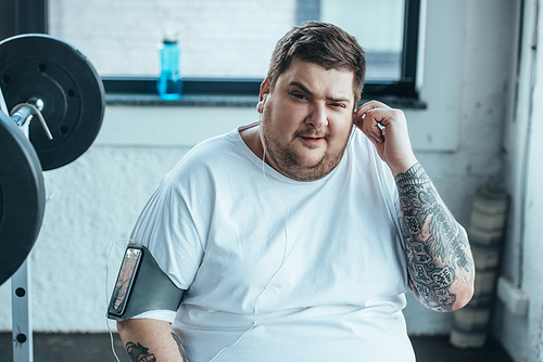 overweight man in smartphone armband  while putting on earphones at gym