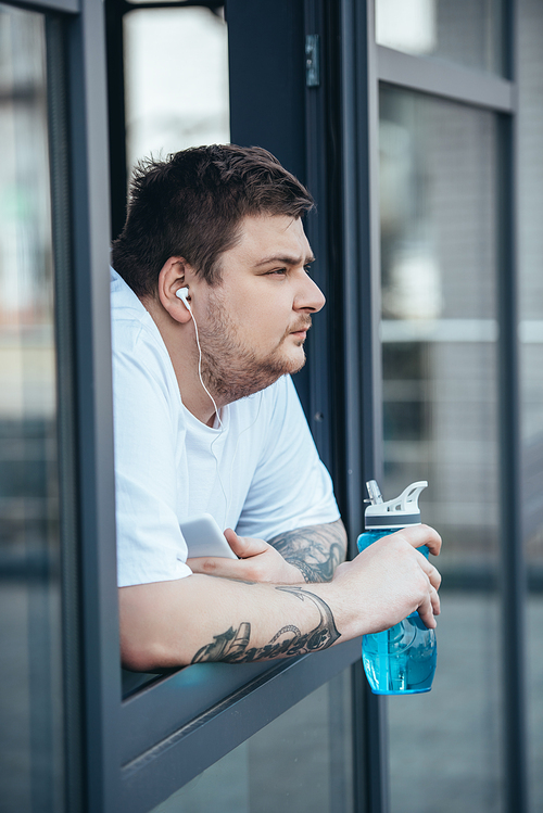 overweight man with earphones and smartphone holding sport bottle and looking out through window at gym