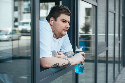 overweight man with earphones and smartphone holding sport bottle and looking out through window at gym