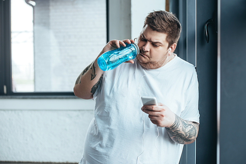overweight tattooed man using smartphone and drinking water from sport bottle at gym