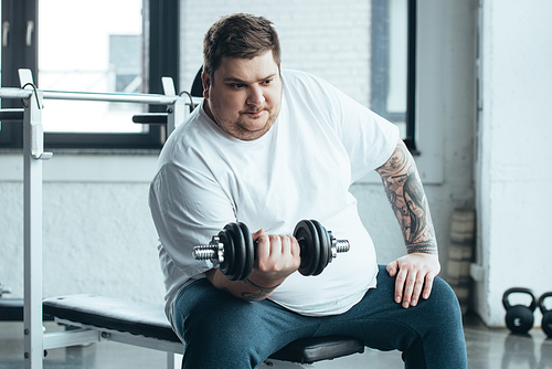 Overweight tattooed man sitting and training with dumbbell at sports center