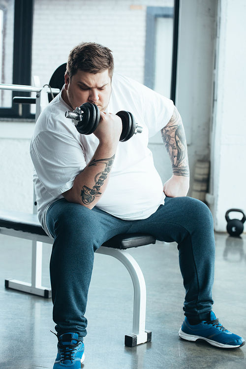Overweight tattooed man sitting and training with dumbbell at sports center