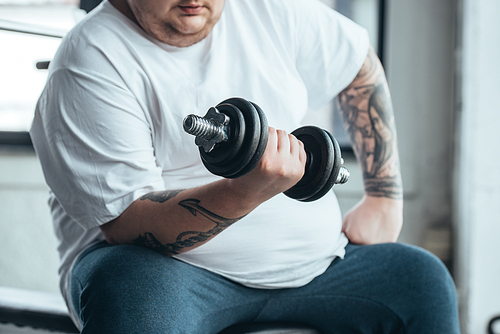 cropped view of Overweight tattooed man sitting and training with dumbbell at gym