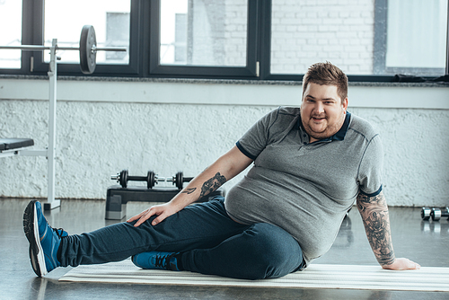 overweight tattooed man , smiling and sitting on fitness mat at sports center