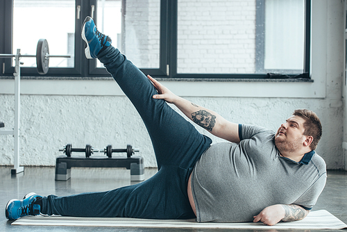 overweight tattooed man lying on fitness mat and stretching legs at gym