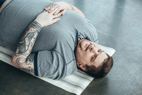 tired overweight tattooed man in grey t-shirt  while lying on fitness mat at sports center
