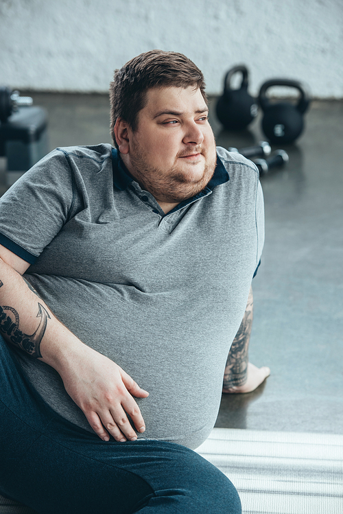 overweight tattooed man looking away while sitting on fitness mat at sports center