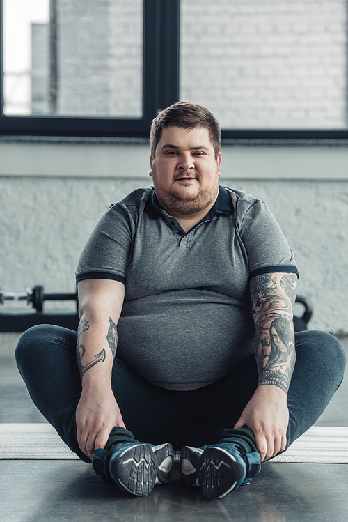 overweight tattooed man sitting and Looking At Camera during stretching exercise at gym