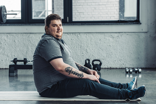 overweight tattooed man sitting on fitness mat and  at sports center