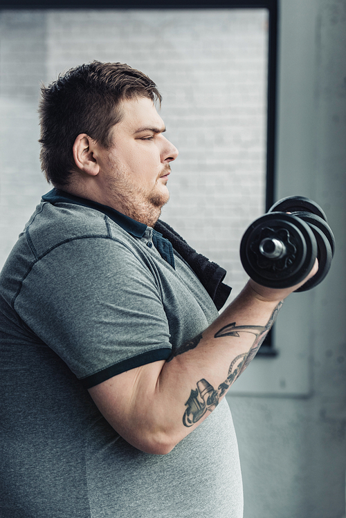 side view of Obese tattooed man exercising with dumbbells at sports center