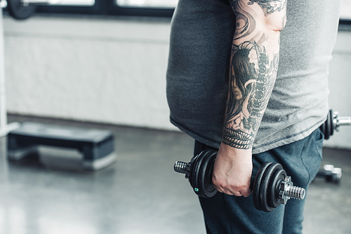 cropped view of Overweight tattooed man training with dumbbells at sports center with copy space
