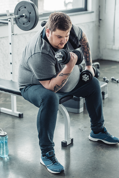 Overweight tattooed man sitting and exercising with dumbbells at sports center