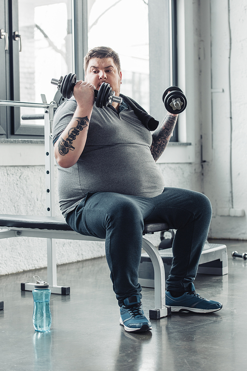Overweight tattooed man sitting and exercising with dumbbells at sports center