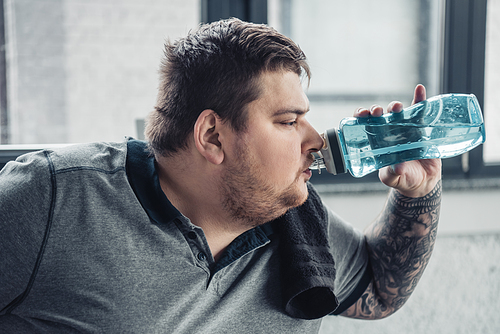 overweight tattooed man drinking water from sport bottle at sports center