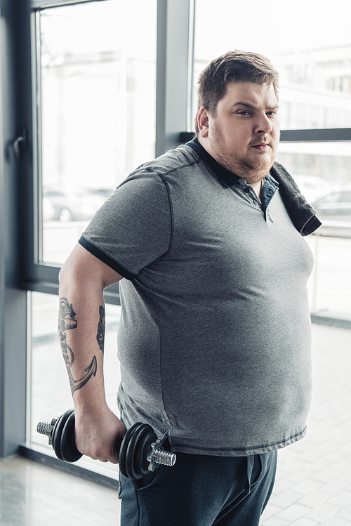 Overweight tattooed man in grey t-shirt with towel exercising with dumbbell at sports center