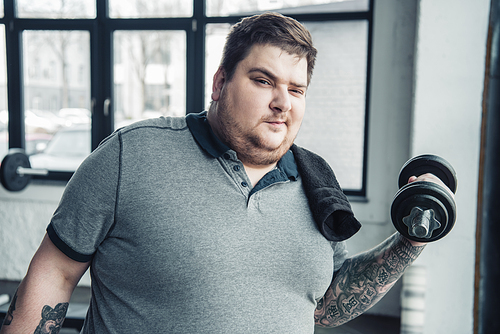 Overweight tattooed man with towel exercising with dumbbell and Looking At Camera at sports center