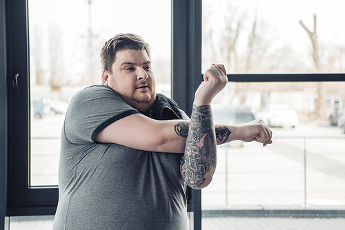 overweight tattooed man in grey t-shirt stretching arms at gym