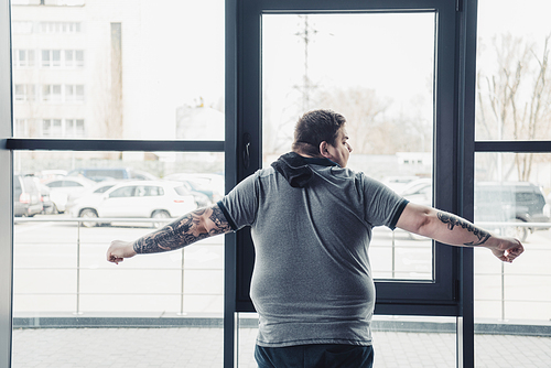 back view of overweight tattooed man in grey t-shirt stretching at gym